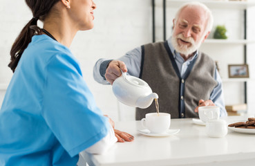 Happy and smiling senior man, pouring tea in white cup, sitting on kitchen at home