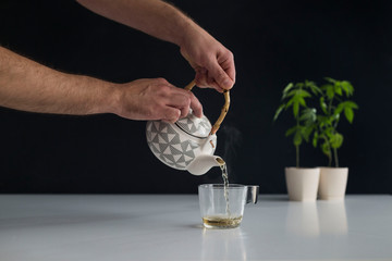 White table with Marijuana plant in pottery. Hand pouring hot CBD tea in to glass.