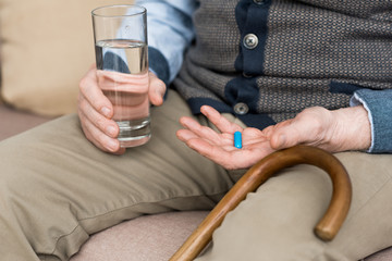 Cropped view of elderly man holding pill and glass with water in hands, sitting on couch