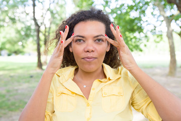 Positive woman touching temples in park. Young lady looking at camera with blurred green trees in background. Nature and concentration concept. Front view.