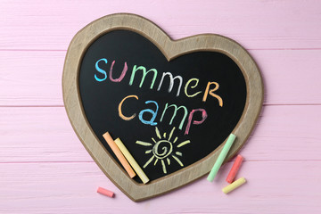Heart shaped blackboard with text SUMMER CAMP, drawing and chalk sticks on wooden background, flat lay