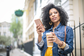 Beautiful girl walking in the street, checking her phone and drinking a coffee