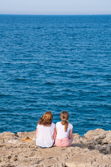 A couple of red hair girls, mother and daughter, sisters or friends standing on the rocks and looking at the blue sea, vertical