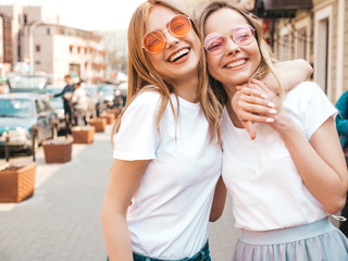 Portrait of two young beautiful blond smiling hipster girls in trendy summer white t-shirt clothes. Sexy carefree women posing on street background. Positive models having fun in sunglasses.Hugging
