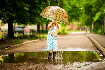 Happy funny kid girl with umbrella jumping on puddles in rubber boots and in polka dot dress and laughing