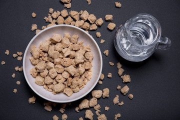 Bol con proteína de soja texturizada fina y gruesa junto a una jarra con agua en fondo negro.