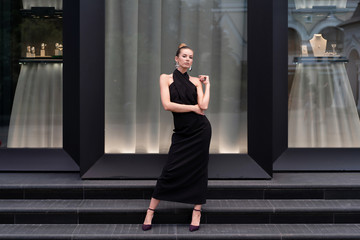 Fototapeta na wymiar Beautiful girl in a long black dress posing in full growth near the boutique. Graceful interesting poses.