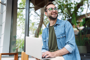 Photo of positive young man typing on laptop while working in city cafe outdoors