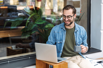 Photo of caucasian young man holding cup of coffee and typing on laptop while working in city cafe outdoors