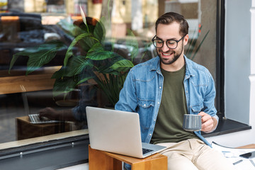 Photo of caucasian young man holding cup of coffee and typing on laptop while working in city cafe...