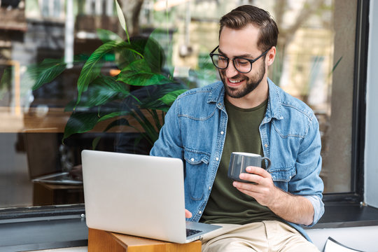 Photo of pleased young man holding cup of coffee and typing on laptop while working in city cafe outdoors