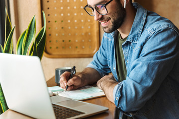 Photo of masculine bearded man writing notes and using laptop while working in cafe indoors