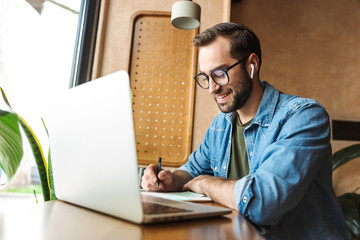 Photo of handsome bearded man writing notes and using laptop while working in cafe indoors