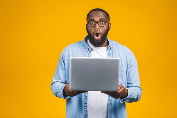 Young surprised african man standing and using laptop computer isolated over yellow background.