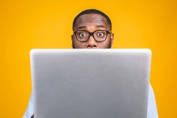 Young surprised african american man standing and using laptop computer isolated over yellow...