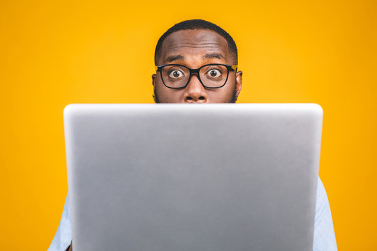 Young surprised african american man standing and using laptop computer isolated over yellow background.