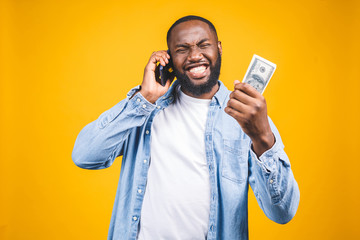 Winner! Young rich african american man in casual t-shirt holding money  dollar bills and mobile phone with surprise isolated over yellow wall.