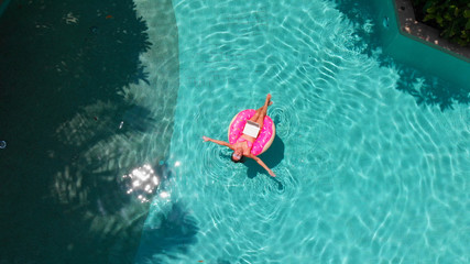 Beautiful young woman with a pink laptop on an inflatable ring in the water in the swimming pool. freelancing, business and travel concept. Aerial view