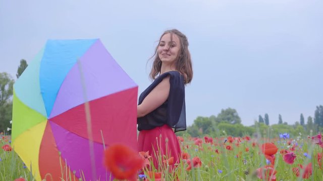 Cute young girl spinning colorful umbrella dancing in a poppy field smiling happily looking in the camera. Connection with nature. Leisure outdoors, summertime fun