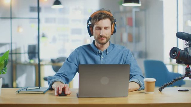 Young Handsome Man Works on a Laptop Computer in Cool Creative Agency in a Loft Office. He has a Take-away Coffee and a Notebook on the Table. He Wears a Jeans Shirt and Headphones.