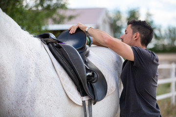 Side view of young guy putting black saddle on back of white horse while standing in paddock on...