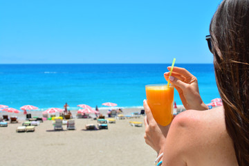 Girl holds orange fresh juice in her hand. Blue sea and beach on a backdrop.
