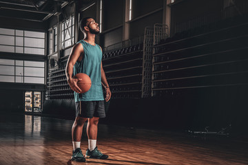 Portrait of Black basketball player holds a ball over a hoop in a basketball hall.