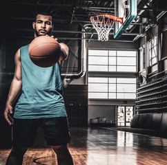Portrait of Black basketball player holds a ball over a hoop in a basketball hall. © Fxquadro