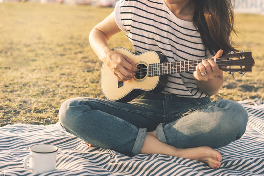 Brunette hipster girl in a striped T-shirt and on a hot day on the beach in soft sunset lighting plays a small guitar ukulele. Relaxed atmosphere, lifestyle image, copy space for text. Fashionable mus
