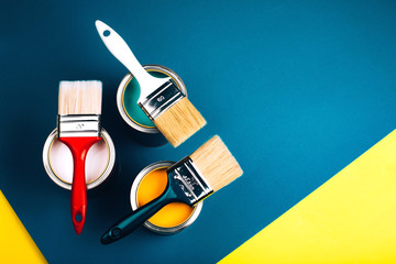Three open cans with brushes on them on yellow and blue background. Yellow, pink and turquoise colors of paint. Top view. Renovation concept.