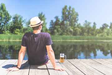 Rear view of hipster man with beer sitting on wooden dock and looking at beautiful lake.