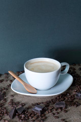 A cup of fresh milky coffee with cream and a wooden spoon on the gunny sack table with coffee beans and chocolate