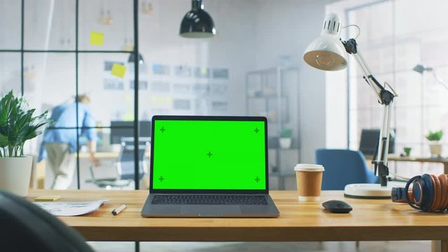 Laptop on the Desk in the Office Shows Green Mock-up Screen. In the Background Bright Modern Open Space Creative Office HUB with Professional Working