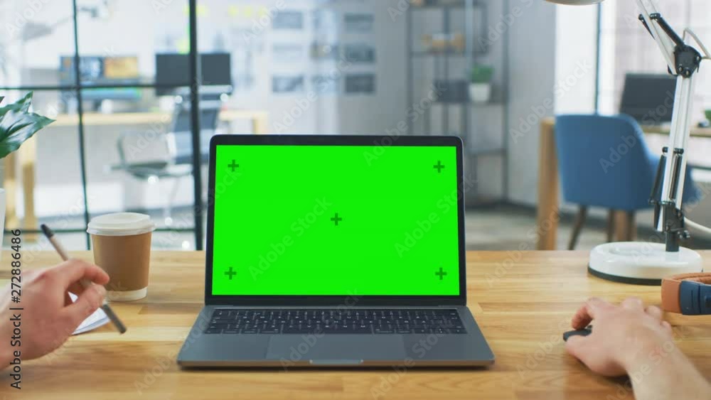 Poster Point of View Shot: Man Uses Laptop Computer with Green Mock-up Screen while Sitting at His Desk. He Holds Mouse and Pen in His Hands. In the Background Stylish Modern Open Space Office
