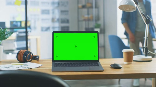 Laptop on the Desk in the Office Shows Green Mock-up Screen. In the Background Bright Modern Open Space Loft Office HUB with Professional Working