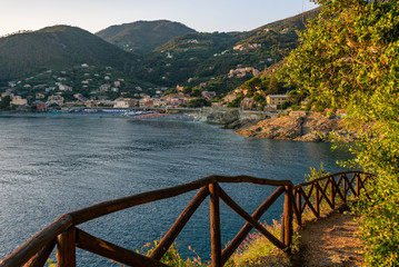 Mountain path secured by a wooden fence, leading to the Mediterranean sea. In the distance, the beach and green hills with colorful houses of Bonassola, Liguria, Italy.