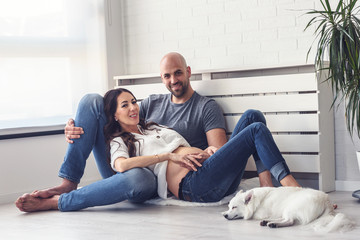 Smiling love couple expecting baby enjoying in their empty apartment with their cute white dog. How to Introduce Dogs and Babies