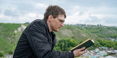 Naklejka na ściany i meble Man reading book in green hilly field. Side view of young handsome man in glasses and warm jacket reading book in open space on background of green hills