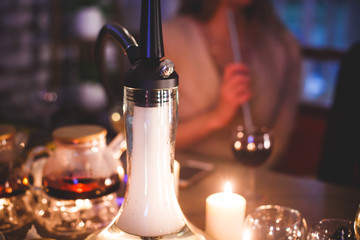 View of Hookah with candles and smoke on a table, hookah lounge with young people smoking in the background, shisha party club concept
