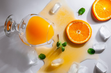 An overturned glass with orange juice lies on a white table. Juice spread over the surface. Around the glass are ice cubes, orange slices, mint. View from above. White background.
