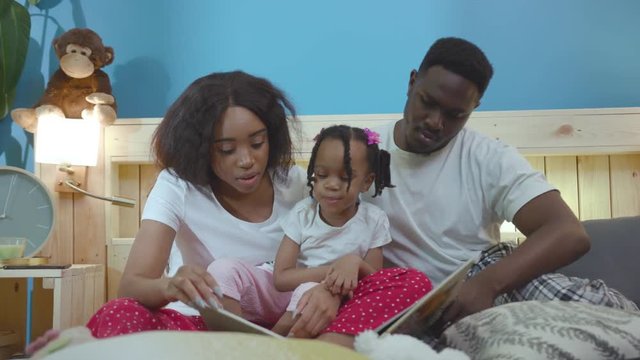 Black African loving parents upbringing a child reading exciting stories in a book lying in bed. Beautiful lifestyle family portrait.