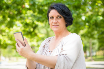 Fototapeta premium Peaceful relaxed woman watching content on phone while waiting someone in park. Middle aged Caucasian lady sitting on bench, holding smartphone and looking away. Online connection concept