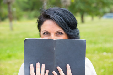 Smiling sly woman covering low face with open notebook with blank cover. Middle aged Caucasian lady reading notes outdoors, hiding face behind copybook and looking at camera. Keeping secret concept