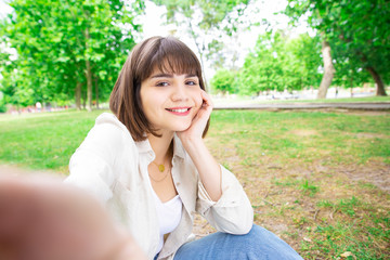 Happy pretty woman taking selfie photo and sitting on lawn. Young lady posing, holding camera which is out of view, sitting on ground with trees in background. Selfie and nature concept. Front view.