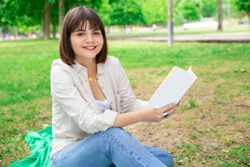 Smiling young woman sitting and reading book on lawn. Pretty lady looking at camera and sitting on blanket on ground with trees in background. Education and nature concept. Front view.