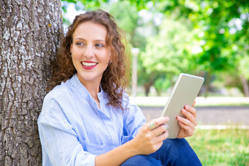 Excited pretty woman enjoying mobile app on tablet. Happy beautiful girl using gadget while resting in park. Internet concept