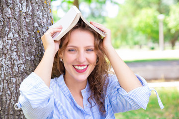 Jolly girl with book above head preparing for exam in park. Portrait of happy young woman in casual...