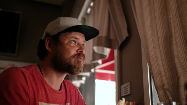 blue-eyed hipster guy with a beard and a cap snapback eating a sandwich (burger)  in a street cafe eating a sandwich (burger) in a street cafe. 4k. 4k video
