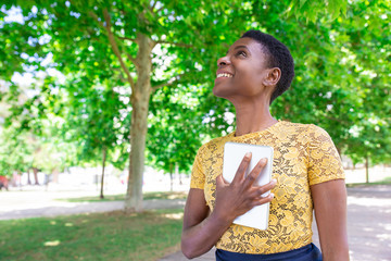 Smiling inspired black woman cuddling tablet to chest in park. Satisfied girl with short hair...