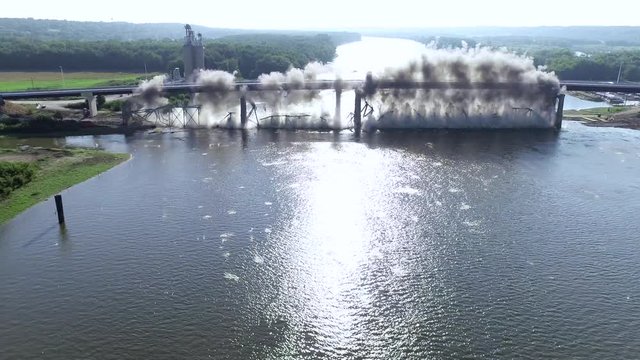 Still aerial shot from high angle of a massive bridge being entirely exploded as a result of controlled demolition. Destroyed construction falls into the river.